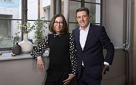 Frau und Mann in formeller Kleidung stehen vor einem Fenster mit Vasen und Pflanzen